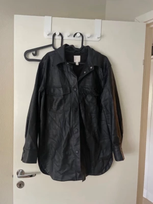Svart skinnjacka från H&M - Stilren svart skinnjacka från H&M med klassisk krage och två stora bröstfickor. Jackan har knäppning framtill och långa ärmar med knapp vid ärmslut. Perfekt för dig som vill ha en trendig och tidlös look. Oversized