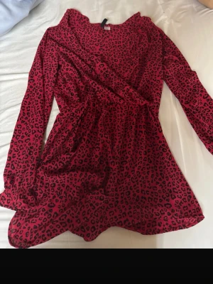 Röd leopardmönstrad omlottklänning - Snygg röd omlottklänning med svart leopardmönster och långa ärmar. Klänningen har en markerad midja och mjukt fallande tyg som ger en feminin siluett. Perfekt för dig som vill sticka ut med ett djurmönster.