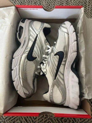 Nike Zoom Vomero 5 vita/silver sneakers - Nike Zoom Vomero 5 sneakers i vitt och silver med svart swoosh. Ovandel i mesh och syntetmaterial för bra andningsförmåga och komfort. Tjock, dämpad sula och klassisk snörning. Perfekta för en sportig och trendig look.