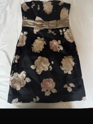 Svart axelbandslös klänning med rosett - Axelbandslös svart klänning med stora blommiga mönster i beige och ljusrosa. Klänningen har en bred satinrosett i midjan som ger en elegant touch. Perfekt för sommarens tillställningar.