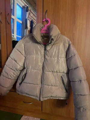 Beige pufferjacka med hög krage - Säljer en beige pufferjacka med hög krage och dragkedja framtill. Jackan har två fickor med dragkedja och quiltad design. Perfekt för kalla vinterdagar och riktigt skön att bära. Slitage på sidan men går att sy, se bild.