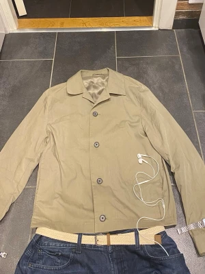 Grisch lätt jacka - Stilren beige overshirt med klassisk krage och knäppning framtill. Overshirten har långa ärmar och raka linjer för en clean look. Tillverkad i ett lätt tyg som passar perfekt för lager-på-lager. Inga synliga loggor eller märken.