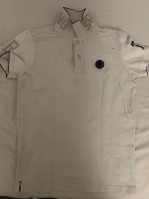 Vit pikétröja från Sail racing - Vit pikétröja från Stone Island med korta ärmar och två knappar framtill. Kragen har tryckt logga och bröstet pryds av det klassiska Stone Island-märket. Perfekt till en avslappnad men snygg stil.