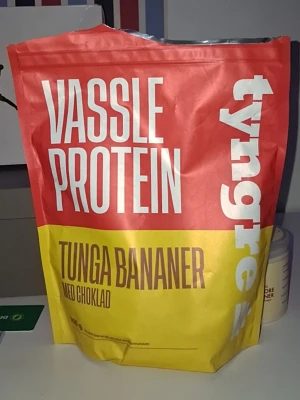 Tyngre vassleprotein tunga bananer - Säljer en påse vassleprotein från Tyngre med smaken tunga bananer och choklad. Förpackningen är röd och gul med tydlig text. Perfekt för dig som vill boosta träningen med god smak. Pulvret är ljust och lätt att blanda ut i vatten eller mjölk.