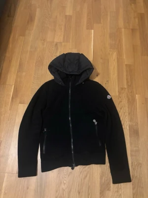 Svart Moncler jacka med huva - Snygg svart jacka från Moncler med stickad struktur och vadderad huva. Jackan har dragkedja framtill, två fickor med dragkedja och Moncler-logga på ärmen. Perfekt för kyliga dagar och ger en stilren look.