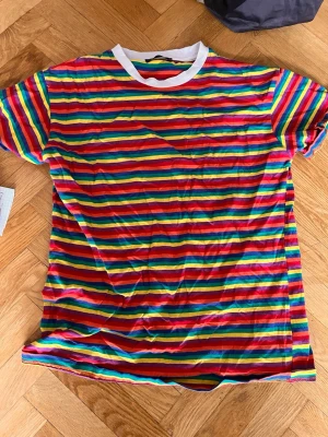 Färgglad randig t-shirt med vit krage - Säljer en färgglad t-shirt med horisontella ränder i rött, blått, grönt, gult och rosa. T-shirten har vit rund halsringning och korta ärmar. Perfekt för dig som gillar färg och vill sticka ut lite extra.Skriv gärna för mått! 💕
