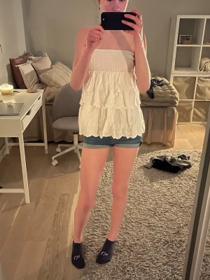 Vit volangig axelbandslös topp/kjol - Säljer en söt vit axelbandslös topp med flera volanglager och smock upptill. Toppen är längre i modellen och har en romantisk känsla, perfekt för varma dagar. Materialet känns lätt och luftigt, och toppen är helt ärmlös för en somrig look. Jag är väldigt osäker på om det är en topp eller en kjol men funkar jättebra som båda, själv tycker jag det är snyggast som en topp!💗🌴