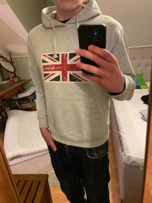 Grå hoodie från Pepe Jeans - Snygg grå hoodie från Pepe Jeans med Storbritanniens flagga på bröstet i nyskick. Klassisk känguruficka framtill, dragsko i huvan och tillverkad i mjukt material. Storlek S 