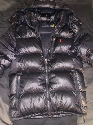 Mörkblå pufferjacka från Polo Ralph Lauren - Snygg svart pufferjacka från Polo Ralph Lauren med blank finish och röd logga på bröstet. Jackan har huva, dragkedja framtill och tre fickor med dragkedja. Perfekt för kalla dagar och riktigt varm tack vare det tjocka materialet.