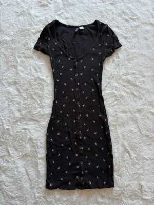 Svart blommig klänning  - Supersöt svart kortklänning med små rosa blommor, knappar framtill och ribbad struktur. Klänningen har korta ärmar och figurnära passform, storlek XXS. 