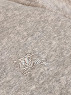 Sko n.2 - Säljer ett grått tygstycke med en broderad logga i vitt och ljusblått. Tyget är mjukt och har en melerad yta. Perfekt för dig som gillar exklusiva detaljer och vill använda det till egna projekt eller dekoration.