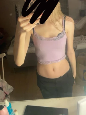 Ljuslila spetslinne crop top - Söt ljuslila crop top med spetsdetaljer i ljusgrått vid halsringning och nederkant. Smala axelband och figurnära passform, perfekt för en trendig och avslappnad stil. Materialet ser ut att vara mjukt och stretchigt.