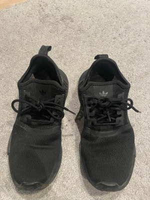 Svarta Adidas sneakers  - Säljer ett par helsvarta Adidas sneakers med mesh-ovandel och klassisk Adidas-logga på plösen. Skorna har svarta snören och en låg siluett som ger en clean och sportig look. Perfekta för dig som gillar stilrena och bekväma sneakers.