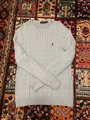 Ljusblå kabelstickad tröja Polo Ralph Lauren - Snygg ljusblå kabelstickad tröja från Polo Ralph Lauren med klassisk rund halsringning och svart broderad logga på bröstet. Tröjan har ribbade muddar vid ärmslut och nederkant. Perfekt för dig som gillar stilrena och tidlösa plagg.