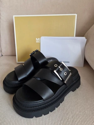 Chunky svarta sandaler Michael Kors - Säljer ett par chunky svarta sandaler från Michael Kors med breda remmar i läder och silverfärgade spännen. Sandalerna har en rejäl platåsula och cool design som passar perfekt till sommarens outfits.