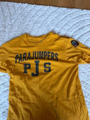 Gul Parajumpers t-shirt med tryck - Gul t-shirt från Parajumpers med stort svart tryck 'PARAJUMPERS PJS' på bröstet och patch på ena ärmen. Klassisk rund hals och normal passform. Perfekt för dig som vill sticka ut med en färgglad och snygg tee. Man får med en gratis vara som tex glasögon sten armband eller bälten helt gratis🔥🤯🤯🔥