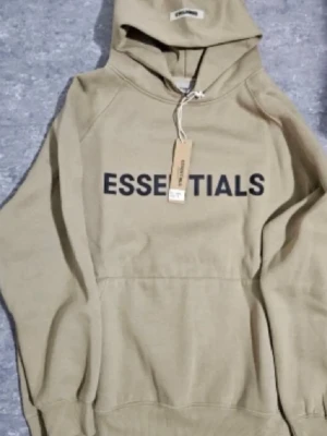 Beige Essentials hoodie med tryck storlek S - Snygg beige hoodie från Essentials med stor svart logga på bröstet. Klassisk känguruficka framtill och huva med dragsko. Tillverkad i mjukt bomullsmaterial som känns skönt mot huden. Perfekt för en avslappnad och trendig streetwear-look.