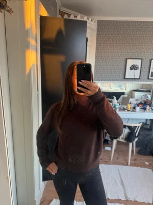 Mörkbrun stickad tröja med polokrage - Mysig mörkbrun stickad tröja med hög polokrage och långa ärmar. Tröjan har en avslappnad passform och ribbstickade muddar vid ärmslut och nederkant. Perfekt för kalla dagar och enkel att matcha med olika outfits.