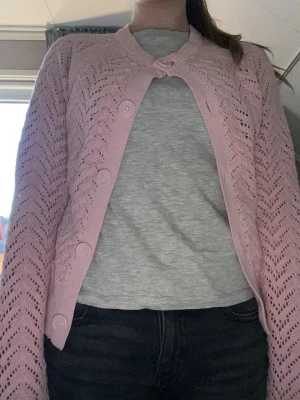 Ljusrosa virkad kofta med knappar - Söt ljusrosa kofta med virkat mönster och stora matchande knappar framtill. Koftan har lång ärm, ribbade muddar och en något kortare passform. Perfekt för lager på lager och ger en romantisk touch till din outfit.