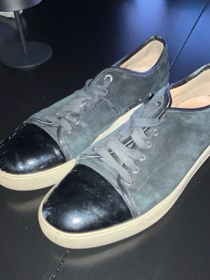 Svarta lanvin cap toe  - Snygga sneakers i svart mocka (bild tagen med blixt så ser grå ut) med svart lackad tå. Fint skick och 100% äkta. Dom är i storlek UK 11 eller svensk storlek 45,5. Ända slitningarna är små defekten på mockan på en del ställen. Skriv för frågor eller funderingar 