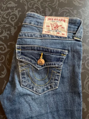 Blå True Religion jeans med kontrastsömmar - Säljer ett par klassiska blå jeans från True Religion med ikoniska kontrastsömmar i gult och snygga detaljer på bakfickan. Jeansen har normal midja, bootcut leg och guldfärgade knappar. 