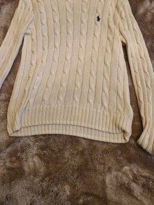 Kabelstickad offwhite tröja Polo Ralph Lauren - Kabelstickad offwhite tröja från Polo Ralph Lauren med klassisk broderad logga på bröstet. Tröjan har rund hals, långa ärmar och ribbade muddar. Perfekt för dig som gillar tidlös och clean stil. Nypris 2300kr har endast defekt i vänstra ärm, inte så märkvärdigt däremot