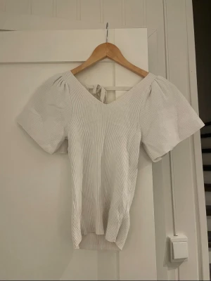 Vit ribbad topp med puffärm och knyt - Superfin vit ribbad topp med v-ringning, korta puffärmar och snygg knytning i ryggen. Perfekt att styla med jeans eller kjol för en trendig look. Toppens material känns mjukt och följsamt.
