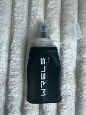 Svart softflaska 150ml - Helt ny aldrig använd. Plombering är kvar. Praktisk svart softflaska med tydliga volymmärkningar upp till 150 ml. Flaskan är tillverkad i mjuk plast och har ett genomskinligt lock och munstycke, perfekt för löpning eller träning. Passar bra i en löparväst eller löparbälte. 