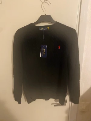 Svart kabelstickad tröja Polo Ralph Lauren - Kabelstickad svart tröja från Polo Ralph Lauren med rund halsringning och den klassiska röda logotypen broderad på bröstet. Tröjan har långa ärmar och ribbade muddar vid ärmslut och nederkant. Perfekt för en stilren och tidlös look.