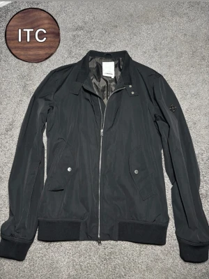 Svart J.Lindeberg jacka  - Snygg svart jacka från J.Lindeberg med dragkedja framtill, två snedställda fickor med tryckknappar och ribbade muddar vid ärmslut och nederkant. Jackan har en diskret logga på ärmen och ett tunt, glansigt innerfoder.