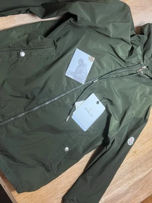 Moncler vindbreaker - Stilren grön vindjacka från Moncler med huva, dragkedja och två fickor framtill. Jackan har Moncler-logga på ärmen och är tillverkad i ett lätt, vattenavvisande material. Perfekt för blåsiga dagar och enkel att matcha med olika outfits. Säljer för att den har blivit förliten. Har tvättas i maskin en gång. 