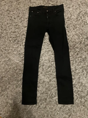 Svarta skinny jeans från Nudie - Säljer ett par svarta skinny jeans från Nudie med klassisk femficksdesign och knappstängning. Jeansen är tighta och har en modern passform som sitter snyggt längs benen. Perfekta för dig som gillar stilrena och enkla jeans.