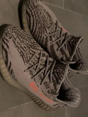 Yeezy Boost 350 V2 Zebra grå - Säljer ett par Yeezy Boost 350 V2 Zebra i grått med svart mönster och orange text på sidan. Skorna har stickad ovandel i Primeknit, rundad tå och ribbad sula i gummi. Klassisk siluett med snörning och bekväm passform.
