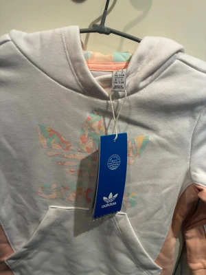 Vit hoodie från Adidas med pastelltryck - Snygg vit hoodie från Adidas med det klassiska Trefoil-loggan i pastellfärger på bröstet. Hoodien har en stor känguruficka framtill, ljusrosa muddar och detaljer samt en mjuk huva med pastellmönstrat innerfoder. Perfekt för en chill och sportig look.
