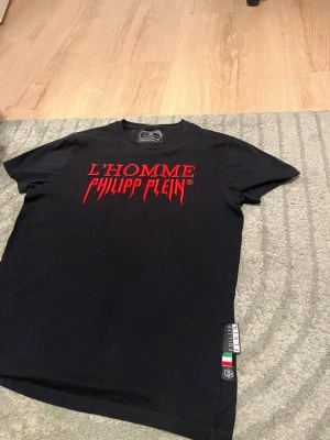 Svart Philipp Plein t-shirt med röd text - Snygg svart t-shirt från Philipp Plein med röd broderad text 'L'HOMME PHILIPP PLEIN' på bröstet. Klassisk passform och rund hals. Liten etikett med italienska flaggan på sidan. Perfekt för dig som gillar streetwear och exklusiva märken.