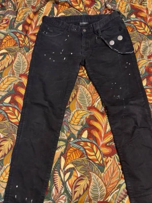 Svarta skinny jeans med kedja armani - Säljer ett par svarta skinny jeans från Emporio Armani med coola vita färgstänk och en avtagbar kedja framtill. Jeansen har fem fickor och en liten logga på bakfickan. Perfekta för dig som gillar en edgy och trendig stil.