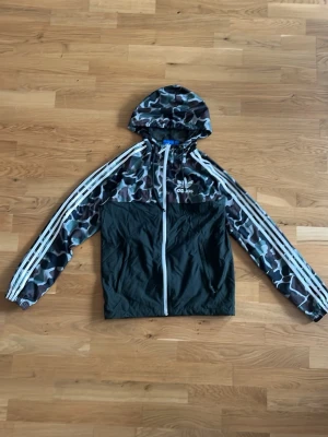 Adidas camo vindjacka med huva - Snygg vindjacka från Adidas med camomönster på huva, axlar och ärmar samt svarta partier på kroppen. Jackan har vita klassiska Adidas-ränder längs ärmarna, dragkedja framtill och två fickor. Tillverkad i lätt syntetmaterial, perfekt för blåsiga dagar. Jätte fint skick vindjackan finns inte längre att få tag i riktigt snygg och skön