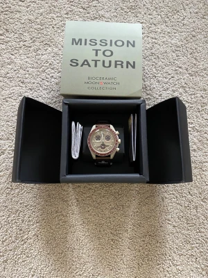 Omega x Swatch Mission to Saturn - Säljer en Omega x Swatch Speedmaster MoonSwatch Mission to Saturn. Klockan har en beige och brun boett i biokeramik, brun textilrem och detaljer i guld och brunt på urtavlan. Kommer i originalbox med tillhörande papper. Perfekt för dig som gillar unika klockor!