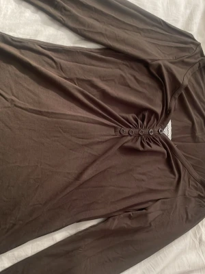 Brun långärmad topp med knappar - Snygg långärmad topp med v-ringning och dekorativa knappar framtill. Tillverkad i mjukt bomullsmaterial som känns skönt mot huden. Perfekt att styla med jeans eller kjol för en trendig look. Ser brun ut men är millitärgröb