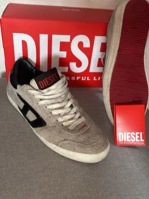 Gråa Diesel sneakers i mocka - Säljer ett par gråa sneakers från Diesel med ovandel i ruggad mocka, svarta detaljer och vit sula. Skorna har klassisk låg profil, vita skosnören och Diesel-logga på plösen. Röd sula undertill och svart D-detalj på sidan ger en unik look. Perfekta skor nu för varmare tider 🙌