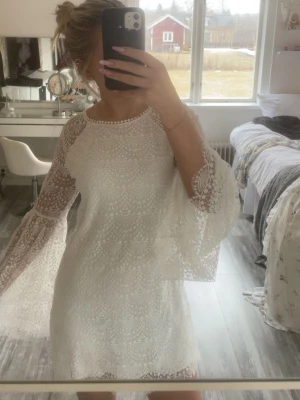 Vit spetsklänning med vida ärmar - Säljer en superfin vit kort spetsklänning med vida, transparenta ärmar och rund halsringning. Klänningen har ett detaljerat spetsmönster och scallop-kant nertill. Perfekt som avslutnings eller stundent klänning.