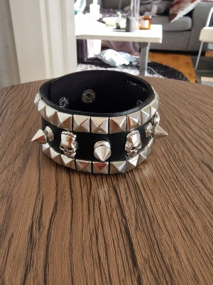 Svart nitar och döskalle armband - Brett svart armband i konstläder med silverfärgade nitar, döskallar och spikes. Armbandet har tryckknappar för justerbar storlek och en cool, rockig stil. Perfekt accessoar för dig som gillar edgy detaljer och vill sticka ut.Skallarmband. PU-läder. 21,5 cm långt.