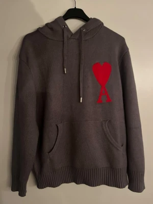 Grå hoodie från Ami Paris med rött hjärta - Säljer en grå stickad hoodie från Ami Paris med stor röd hjärtlogga och bokstaven A på bröstet. Hoodien har dragsko i huvan, känguruficka och ribbade muddar. Perfekt för dig som gillar streetwear och vill ha något som sticker ut.