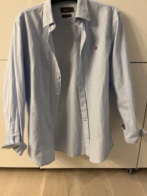 Ljusblå skjorta från Morris - Klassisk ljusblå skjorta från Morris med lång ärm och knappar framtill. Skjortan har en liten röd broderad detalj på bröstet och är tillverkad i mjuk bomull. Perfekt till jeans eller chinos för en clean look.