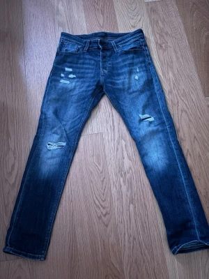Blå slitna jeans från Jack & Jones - Säljer ett par blå jeans från Jack & Jones med slitningar och hål på benen för en trendig look. Jeansen har smal passform, klassiska fem fickor och är gjorda i denim. Perfekta för dig som gillar en avslappnad och cool stil.