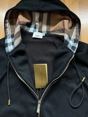 Svart hoodie från Burberry med dragkedja - Svart hoodie från Burberry med klassiskt rutigt foder i huvan och guldfärgade detaljer på dragkedja och snören. Broderad logga på bröstet och mjukt material. Perfekt för dig som vill ha en stilren och exklusiv look.