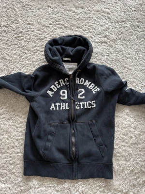 Abercrombie & Fitch hoddie  - Cool  hoodie från Abercrombie & Fitch med vit text 'Abercrombie Athletics 92' på bröstet. Dragkedja framtill, två fickor och skön huva. Perfekt för en avslappnad och sportig stil. Tillverkad i mjukt bomullsmaterial.  De står XXL men sitter som M/L . Skriv vid funderingar eller frågor✌🏼