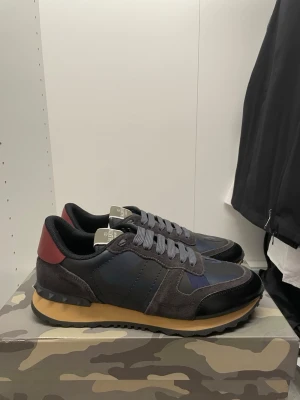 Valentino Rockrunner sneakers mörkgrå - Säljer ett par Valentino Rockrunner sneakers i mörkgrått och svart med röda detaljer på hälen. Skorna har en platt sula i beige och grått, snörning och coola nitar på bakdelen. Materialmix av mocka, skinn och textil. Perfekt för dig som gillar exklusiva sneakers med unik design.