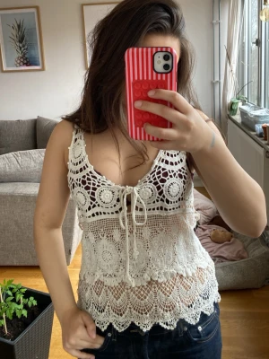 Vit virkad linnetopp med knyt - Supersöt virkad linnetopp i vitt med v-ringning och knyt framtill. Toppen är ärmlös och har ett detaljerat spetsmönster som ger en bohemisk vibe. Perfekt till sommaren och lätt att styla med jeans eller kjol.