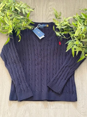 Mörkblå kabelstickad tröja Polo Ralph Lauren - Mörkblå kabelstickad tröja från Polo Ralph Lauren med V-ringning och röd broderad logga på bröstet. Tröjan har ribbade muddar och klassisk passform. Perfekt för lager-på-lager och snygg till jeans. Kvittot finns vid behov fråga gärna om bilder eller info